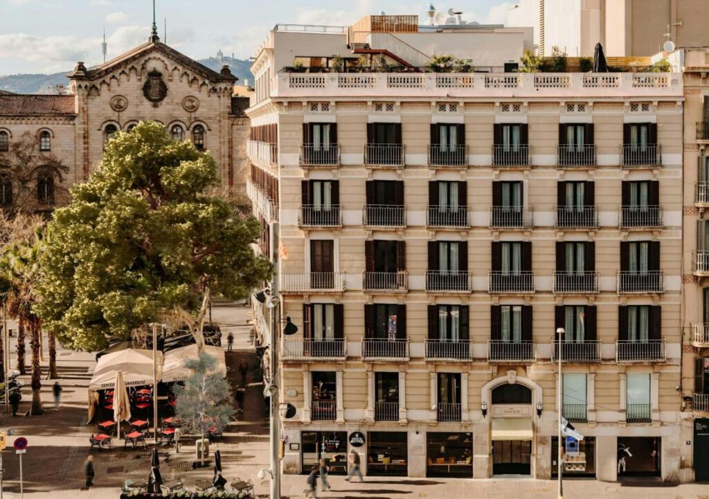 View of an elegant building in Barcelona, featuring balconies and a vibrant street scene with greenery and cafes.