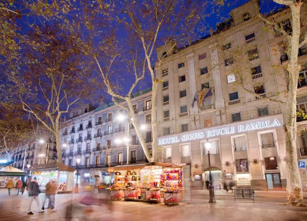 Hotel Serhs Rivoli Rambla illuminated at night, with bustling street vendors and tree-lined promenade in Barcelona.
