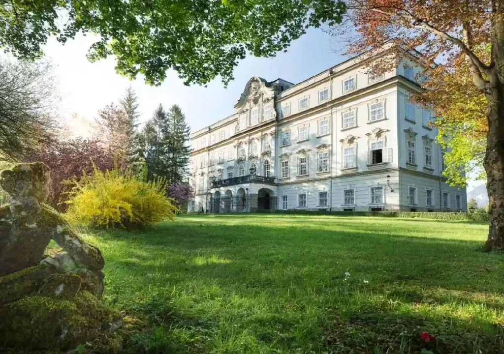 Elegant white mansion surrounded by vibrant greenery and flower gardens under a clear blue sky. Beautiful architecture.