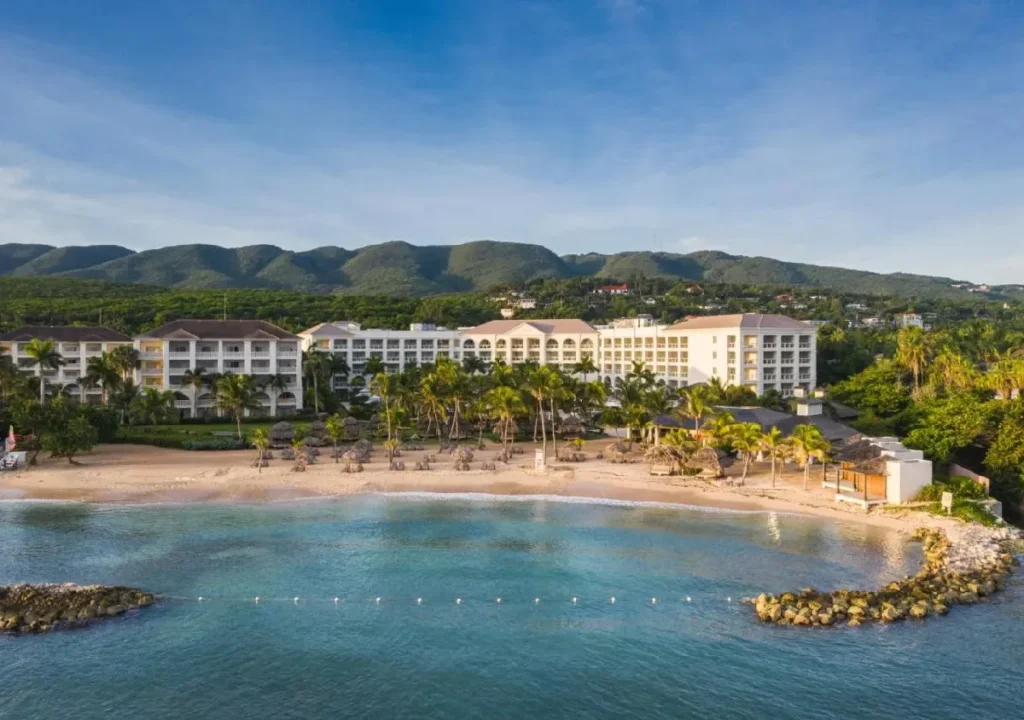 Aerial view of a beachfront resort surrounded by palm trees and mountains under a clear blue sky. Perfect vacation spot.