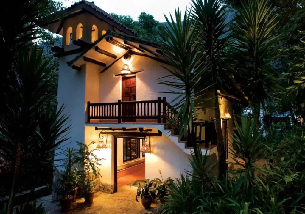 Charming two-story white house with wooden accents, surrounded by lush greenery and warm outdoor lighting. Perfect retreat!