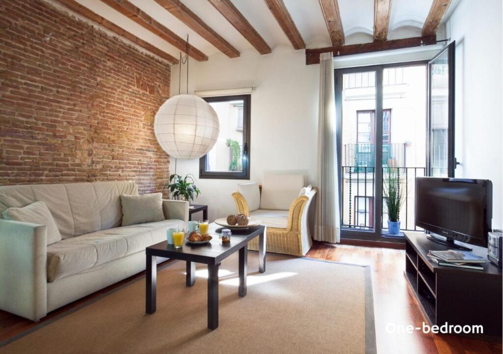 Bright one-bedroom living room featuring rustic brick walls, cozy furniture, and natural light from large windows.
