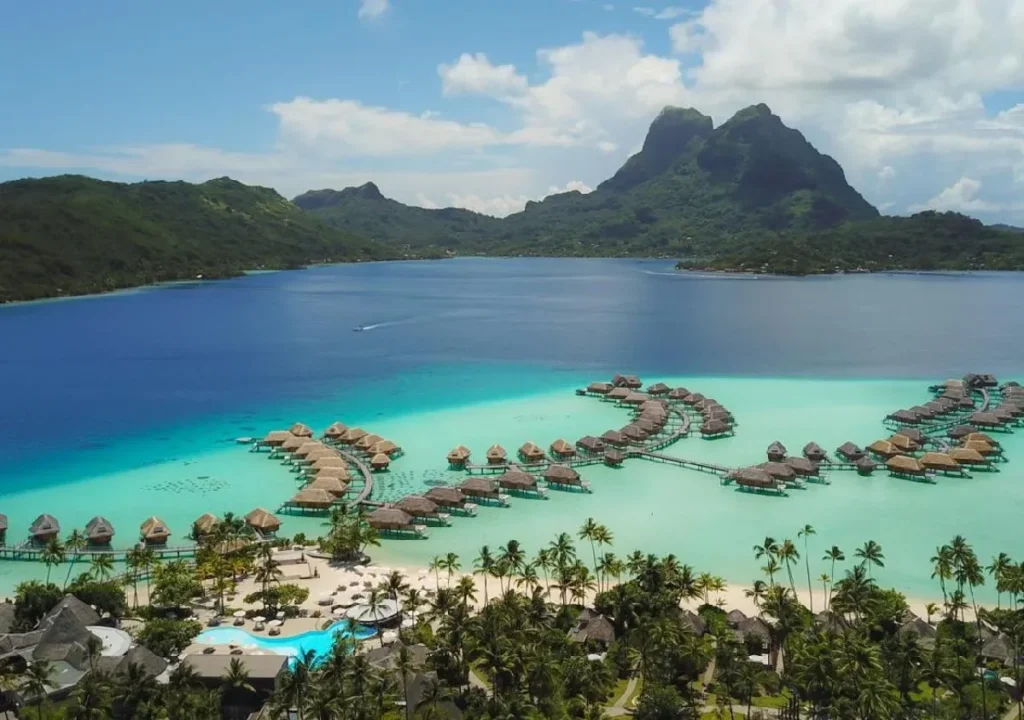 Aerial view of overwater bungalows in turquoise waters, surrounded by lush mountains and palm trees in a tropical paradise.
