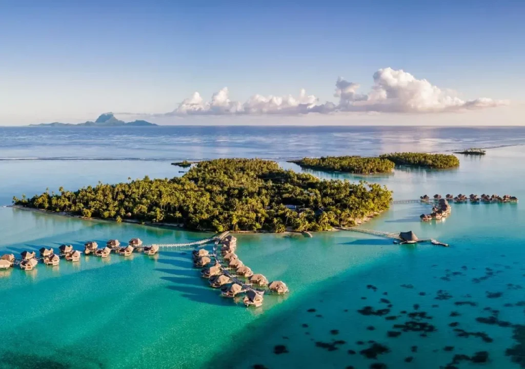 Aerial view of a tropical island with overwater bungalows surrounded by clear turquoise waters and lush greenery.