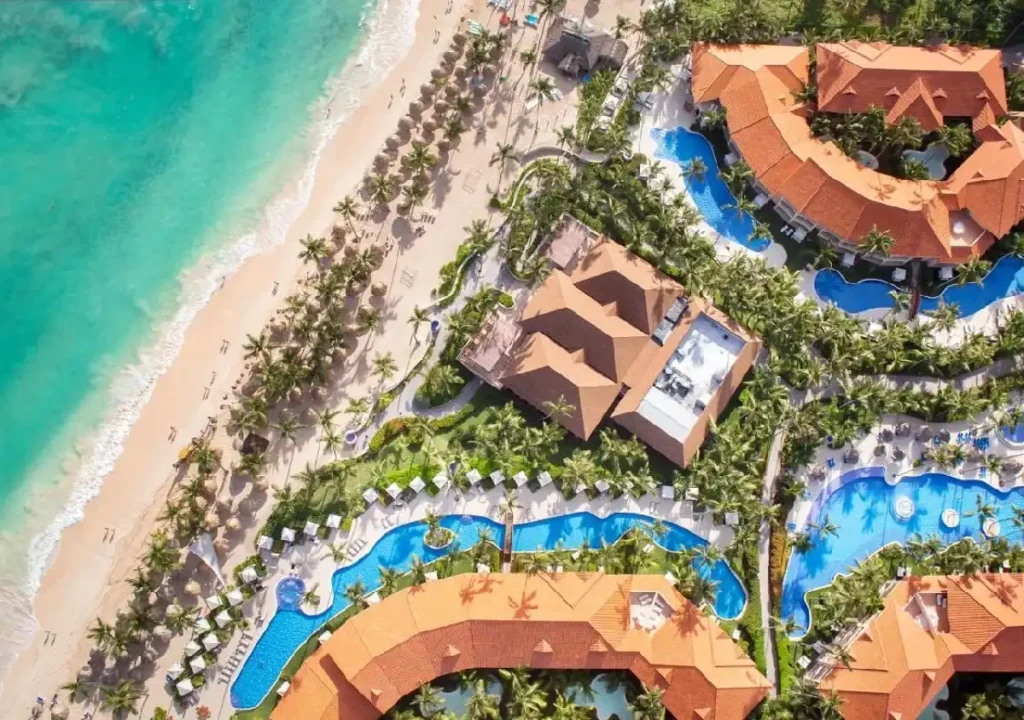 Aerial view of a tropical resort featuring pools, palm trees, and a sandy beach along a vibrant turquoise ocean.