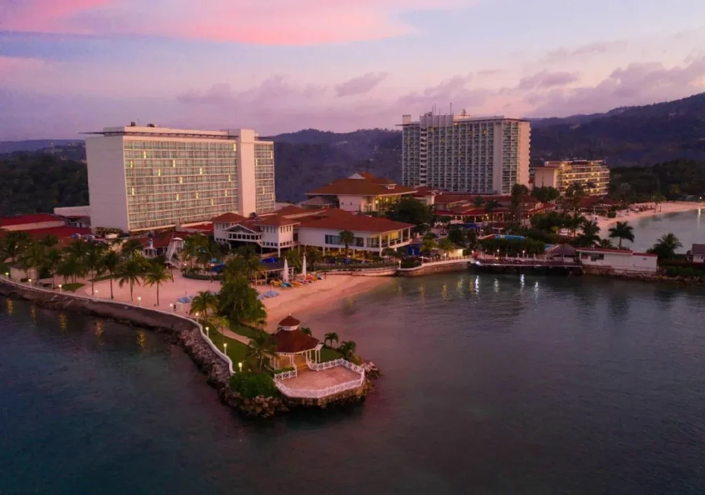 Aerial view of a tropical resort at sunset, featuring lush palms, sandy beach, and stunning coastal scenery.