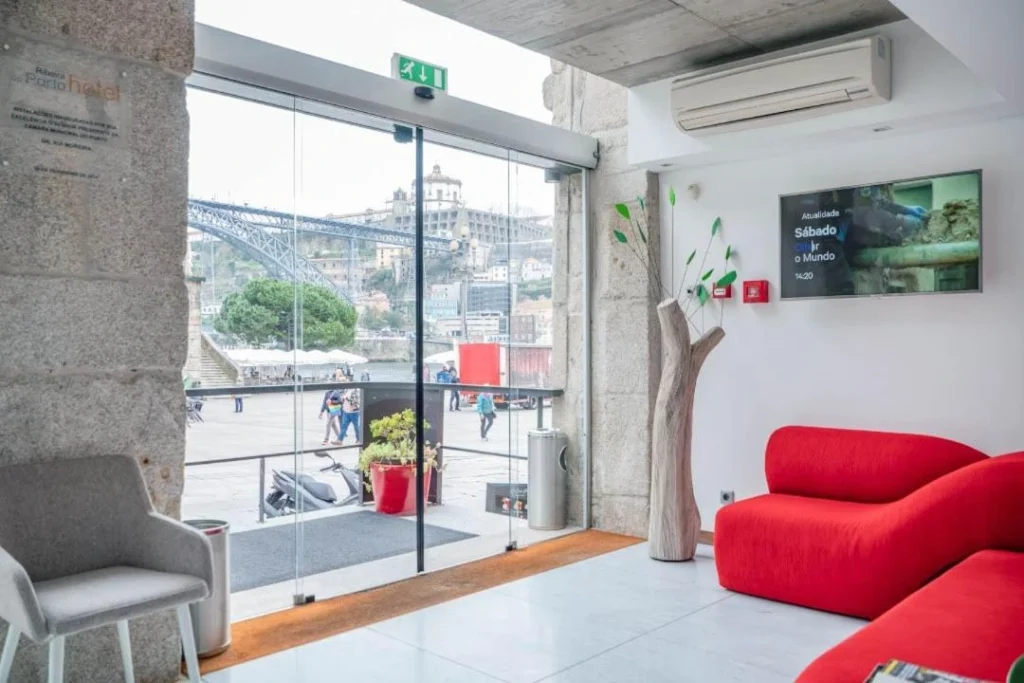 Modern hotel lobby with stylish red seating, glass doors, and a view of the cityscape, featuring greenery and a TV.