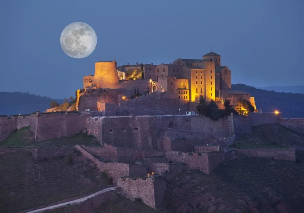 A historic castle illuminated at night with a full moon in the sky, set against a scenic mountainous backdrop.