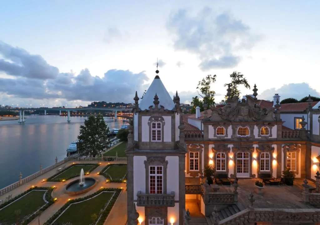 Elegant riverside villa at dusk, showcasing ornate architecture, lush gardens, and scenic views of the water and bridge.