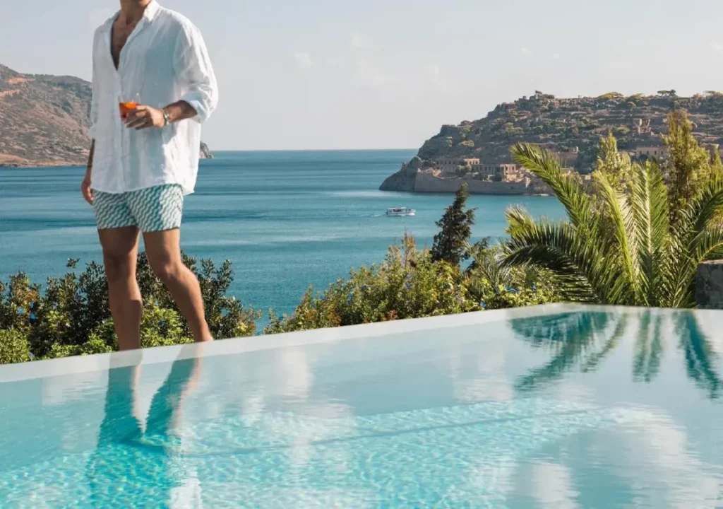 A man in a white shirt and swim shorts enjoys a drink by a serene infinity pool overlooking the ocean.