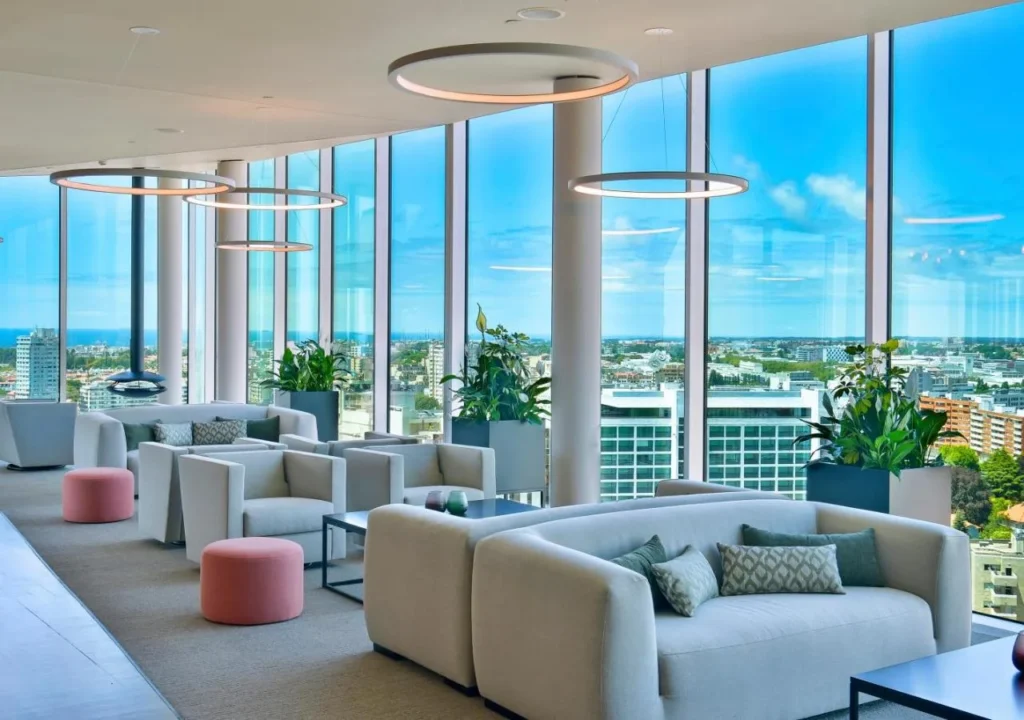 Modern office lounge with plush seating, large windows showcasing a city view, and stylish lighting fixtures.