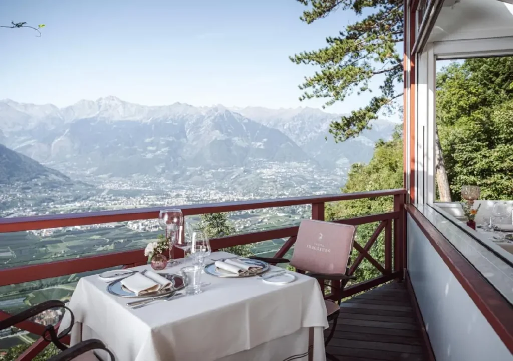 Scenic mountain view from a restaurant terrace, elegantly set table with glasses and a flower arrangement.