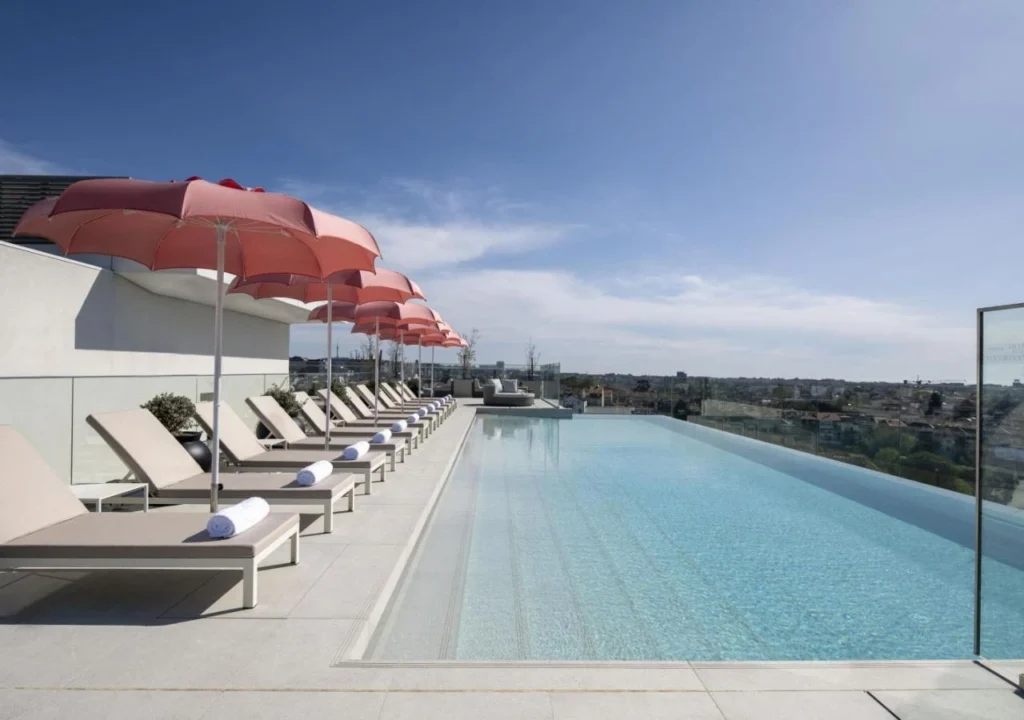 Rooftop infinity pool with loungers and pink umbrellas, offering stunning city views under a clear blue sky.