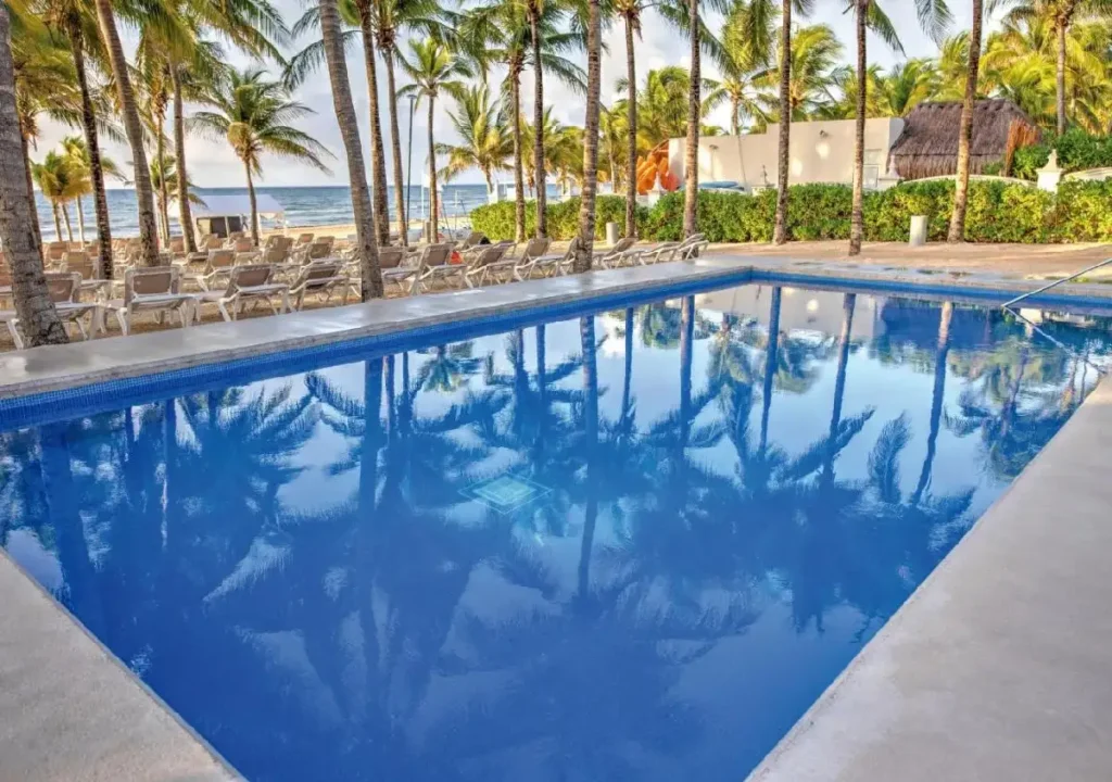 Serene pool reflecting palm trees and beachside lounge chairs under a bright sky.