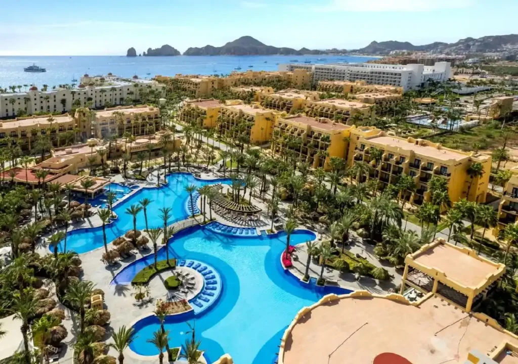 Aerial view of a luxurious resort with palm trees and a beautiful blue pool, overlooking the ocean in Cabo San Lucas.