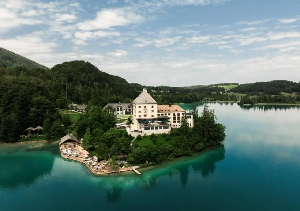 Aerial view of a picturesque hotel on a serene lake, surrounded by lush greenery and mountains under a blue sky.