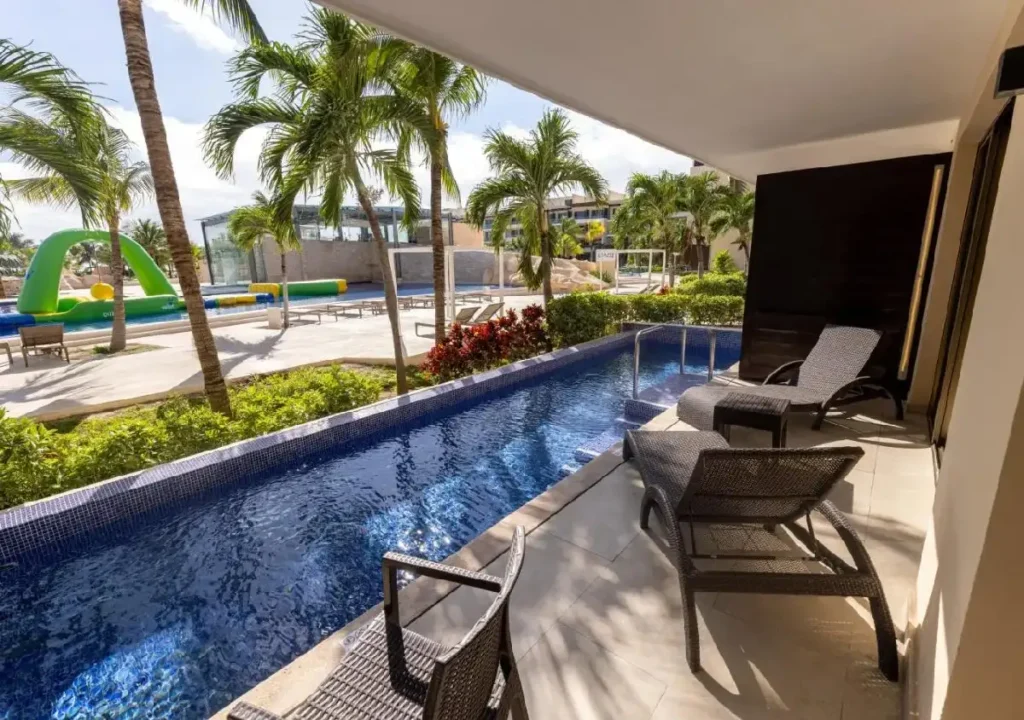 Serene balcony view featuring poolside loungers and a glistening pool surrounded by lush greenery and palm trees.