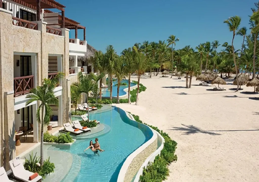 Serene beach resort scene with a pool, sun loungers, and palm trees under a clear blue sky.