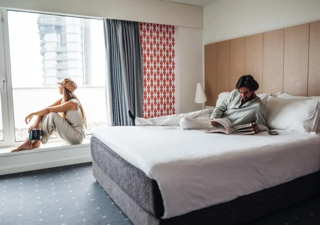 A young woman sits by the window, while a man reads a book on a bed in a stylish hotel room.