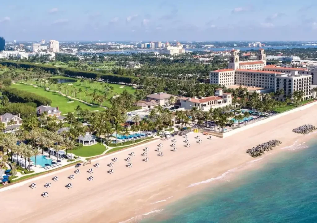 Aerial view of a luxury beach resort with palm trees, sandy beach, and ocean, surrounded by city skyline and greenery.