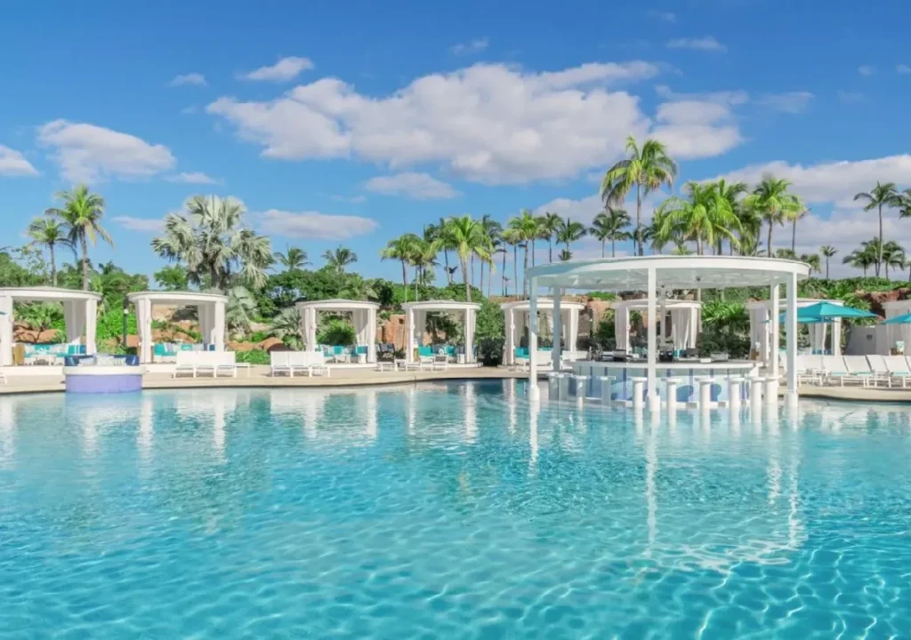 Tropical pool oasis with cabanas, palm trees, and a bar under a blue sky, perfect for relaxation and leisure.