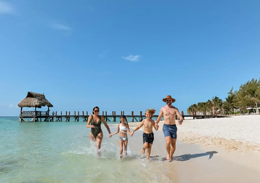 Family running together on a sunny beach, splashing in the water near a wooden pier and tropical palm trees.