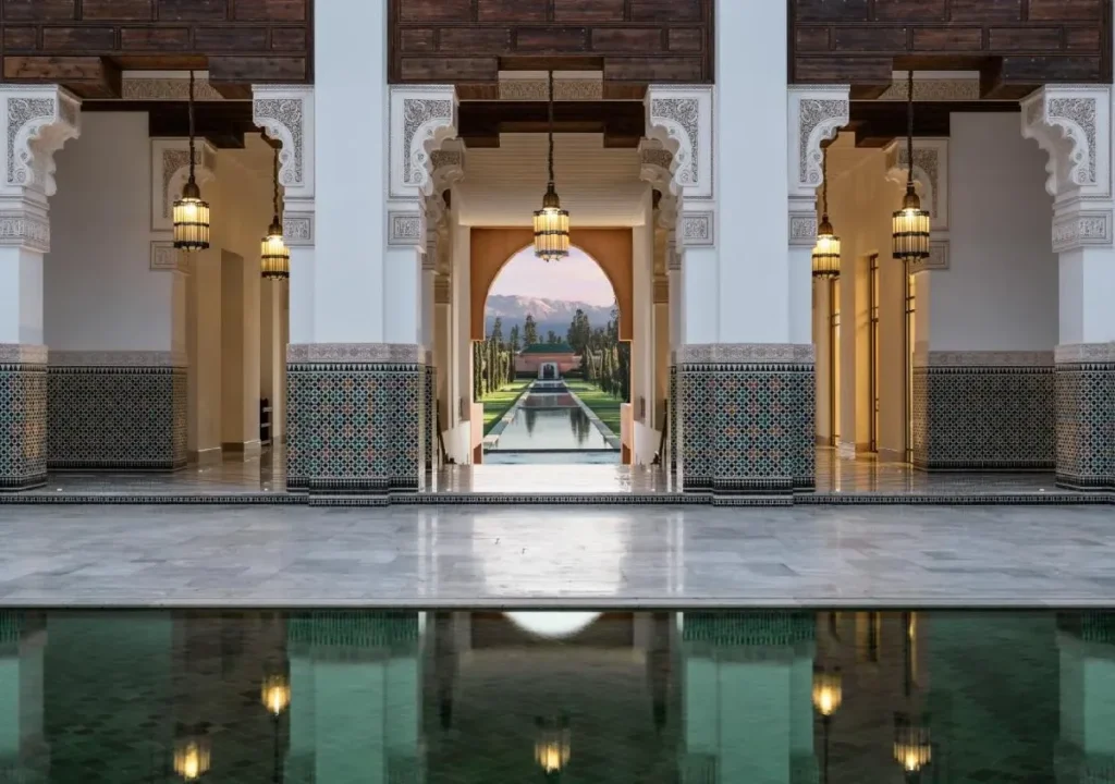 Stunning view of a Moroccan palace interior with reflective pool, arches, ornate decor, and a lush garden pathway.