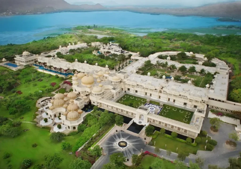 Aerial view of a luxurious resort with intricate architecture, gardens, and a scenic lake in the background.