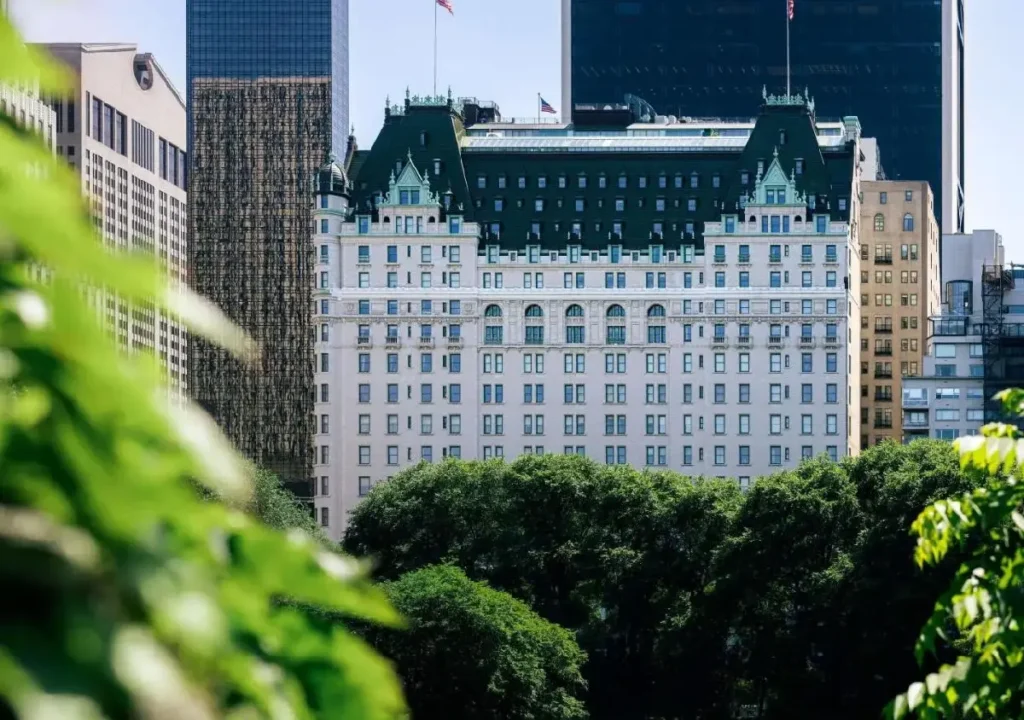 View of a historic hotel surrounded by modern skyscrapers and lush greenery in an urban landscape.