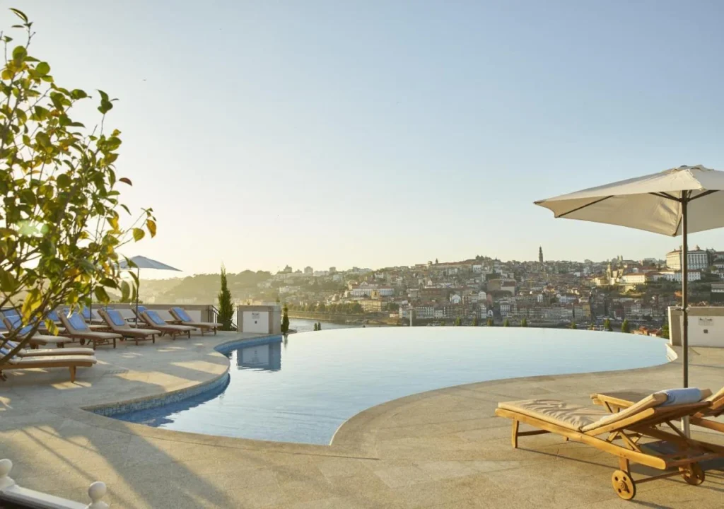 Rooftop infinity pool with city view, lounge chairs, and umbrellas under a clear sky at sunset, ideal for relaxation.