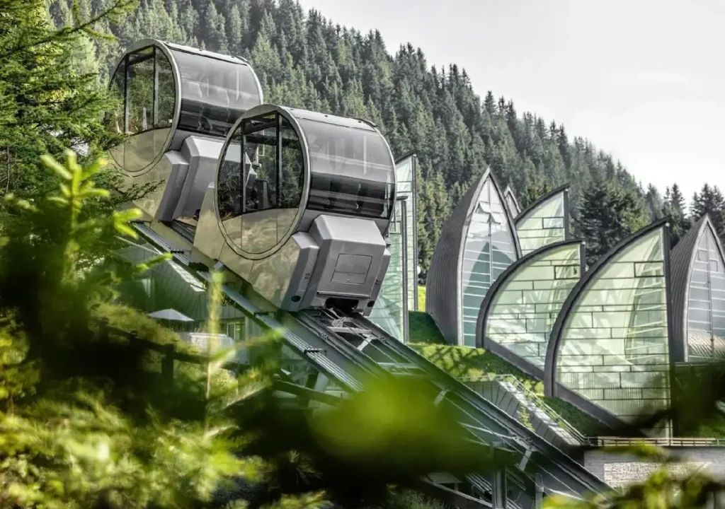 Modern gondolas ascend a hill with lush greenery and unique architectural structures in the background. Scenic mountain view.