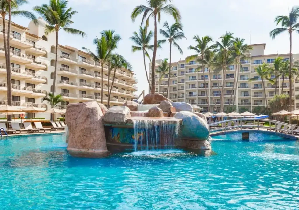 Tranquil resort pool with a waterfall feature, palm trees, and lounge chairs under blue skies. Perfect vacation spot.