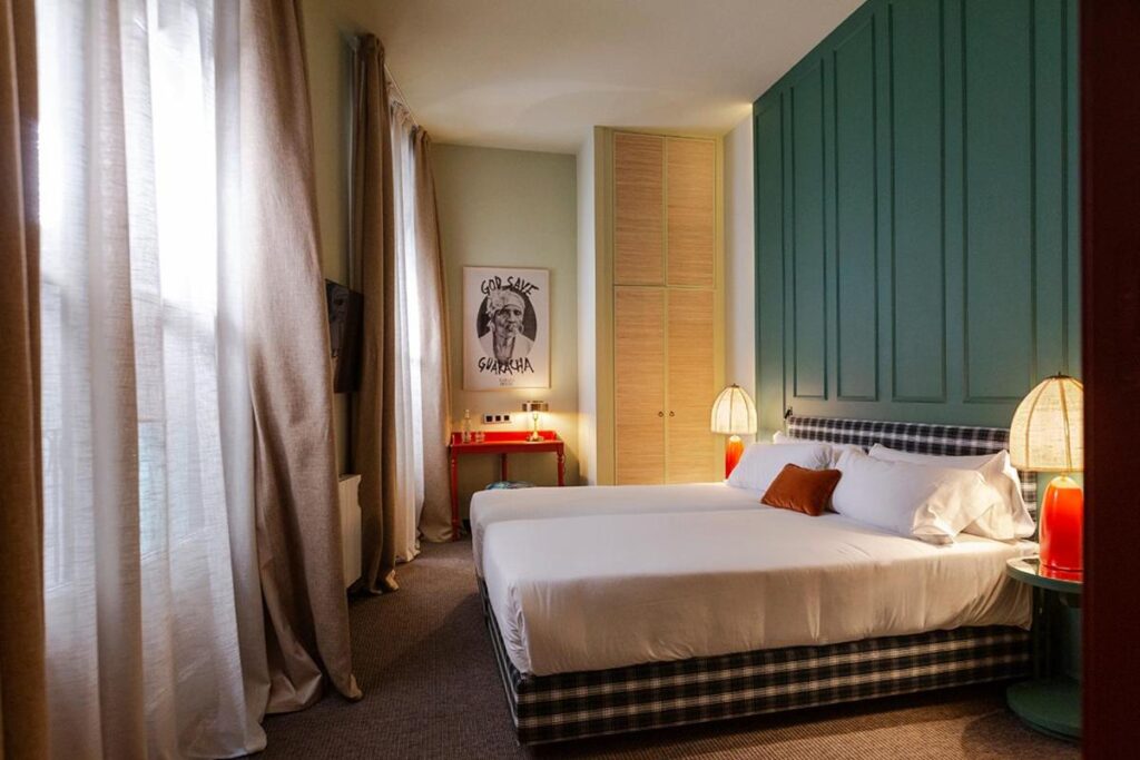 Cozy hotel room with a green accent wall, large windows, and stylish decor featuring a double bed and modern lamps.