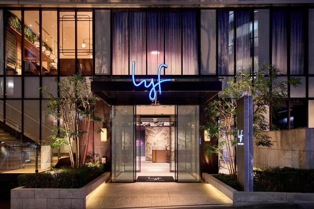 Modern entrance of the lyf hotel featuring a glowing blue sign and sleek glass facade, surrounded by greenery.