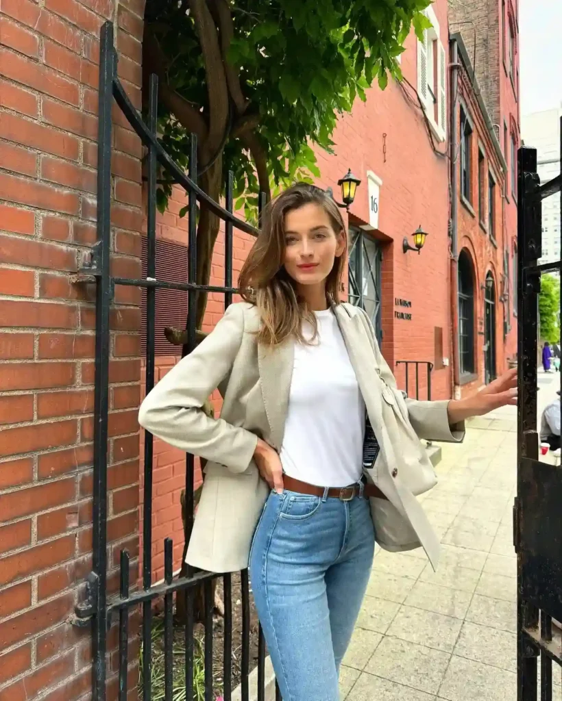 Woman in a stylish blazer and jeans poses near a brick wall and greenery, showcasing a modern urban outfit.