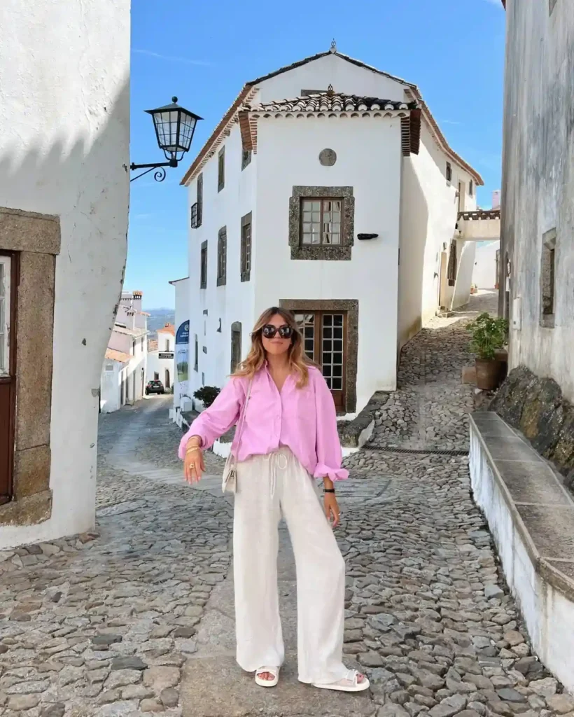 A stylish woman in a pink shirt and white pants stands in a charming cobblestone street among white buildings.