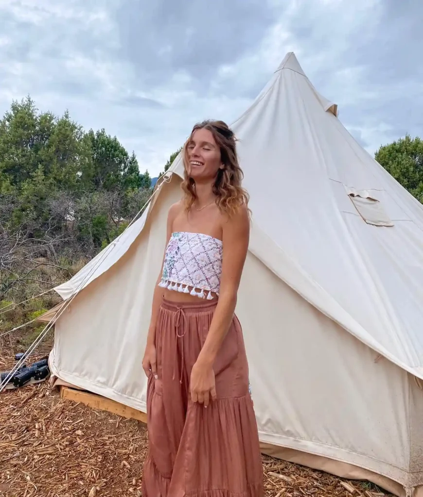 Young woman smiling in a trendy outfit, standing outside a teepee tent in a natural setting with trees and cloudy sky.