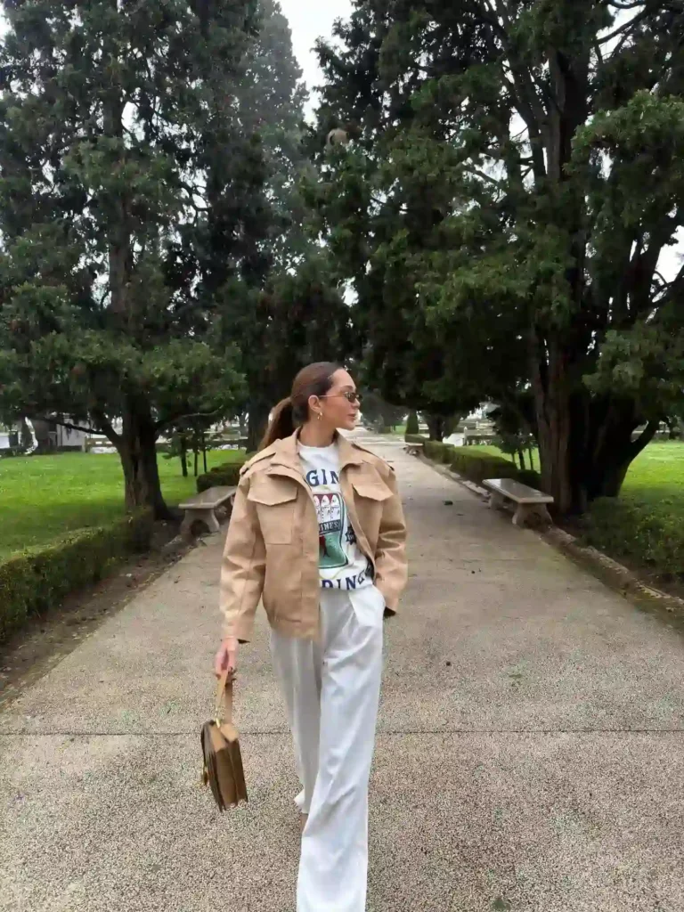 Woman walking in a park, wearing a beige jacket and white pants, holding a brown bag against a backdrop of green trees.