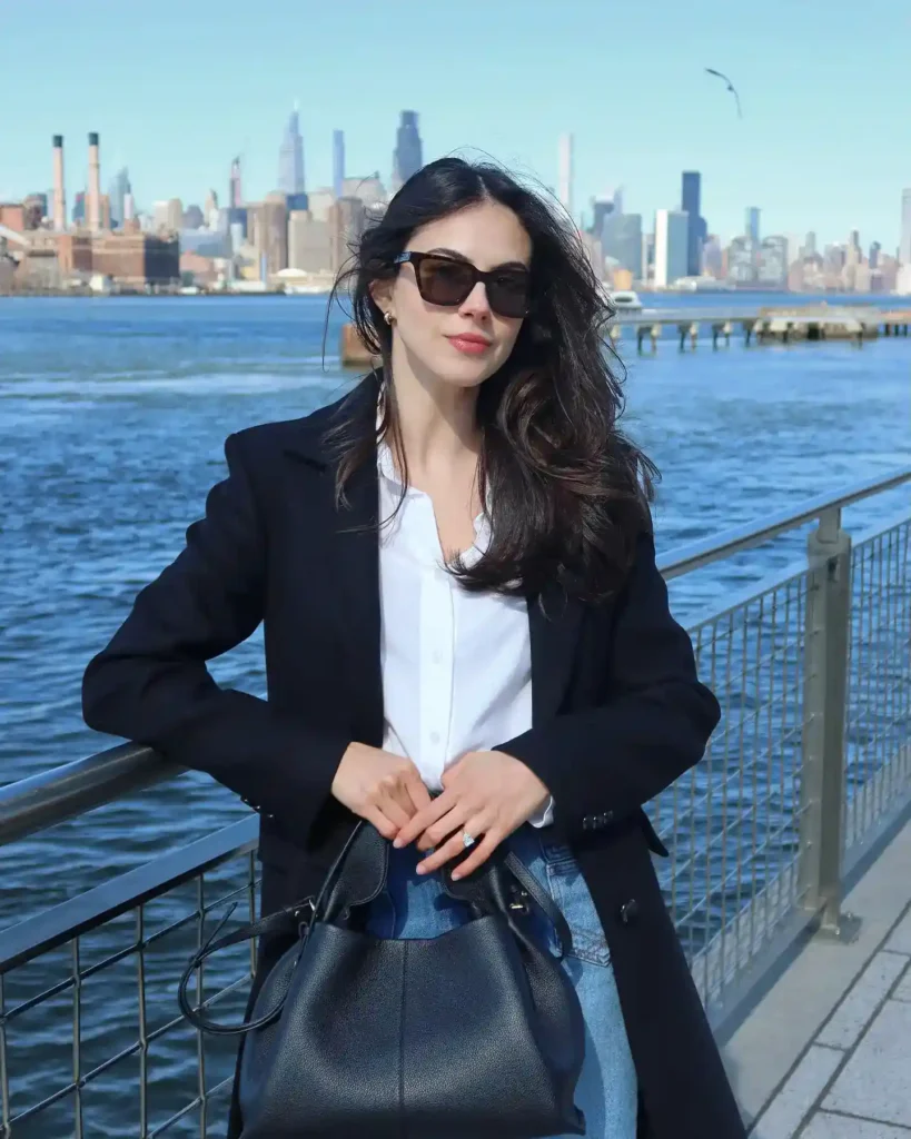 Stylish woman with sunglasses poses near water with a city skyline in the background, showcasing urban fashion.