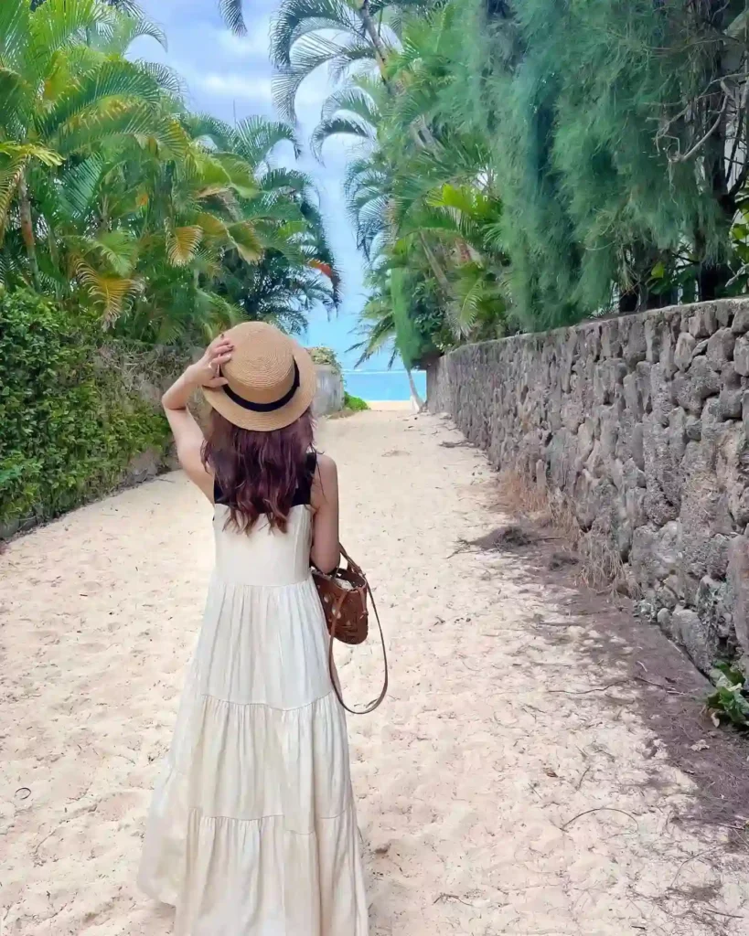 Woman in a flowing white dress and straw hat walks along a sandy path lined with palm trees, heading towards the beach.