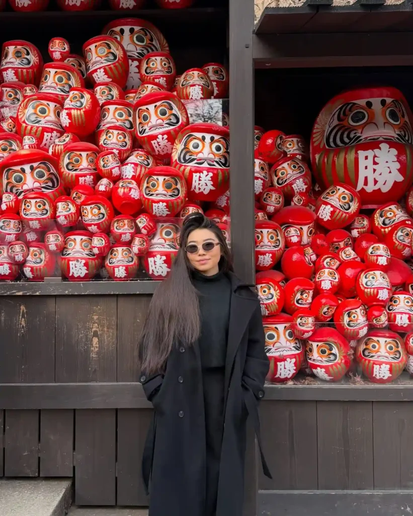 Young woman in sunglasses poses beside a vibrant display of daruma dolls, symbolizing good luck and perseverance.