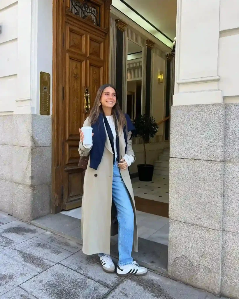 Young woman sipping coffee at a stylish entrance, dressed in a chic trench coat and casual jeans.