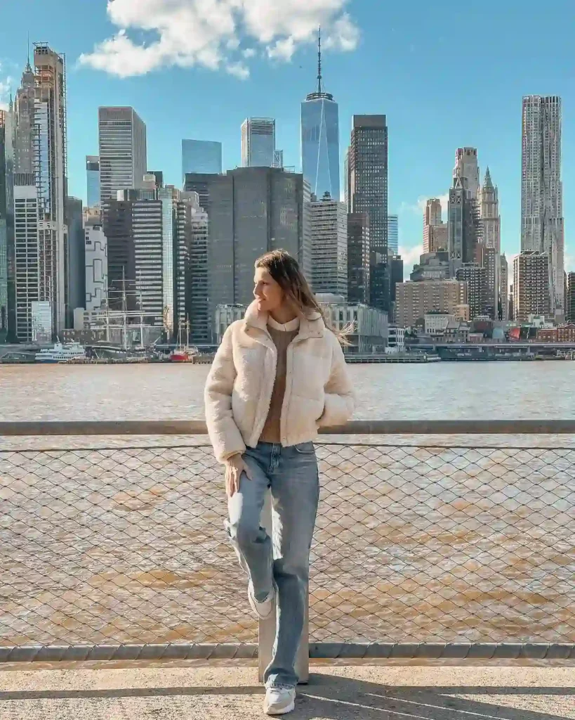 Woman in a cozy jacket poses by the waterfront, with a stunning skyline backdrop in bright daylight.