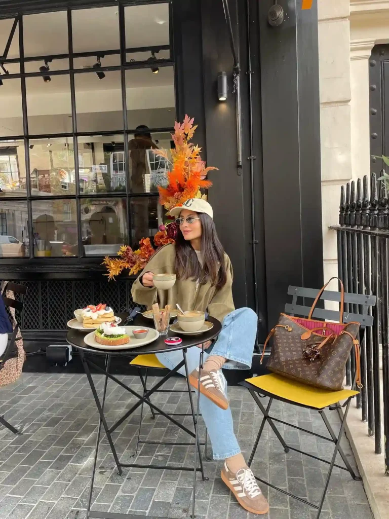 Woman enjoying a cozy café setting with coffee and pastries, surrounded by autumn decorations and stylish accessories.