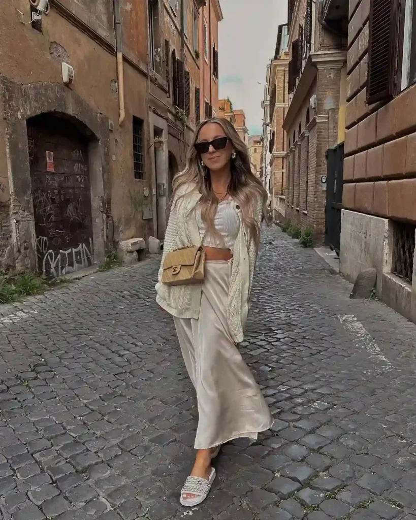 Fashionable woman in sunglasses strolls down a cobblestone street in Rome, wearing a chic outfit with a stylish bag.