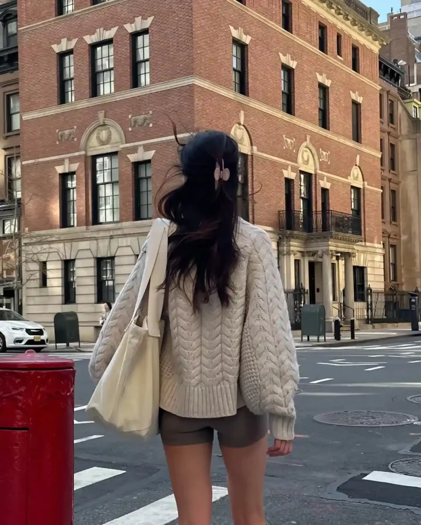 Woman in a cozy sweater and shorts stands on a city street, facing a historic brick building with street elements.