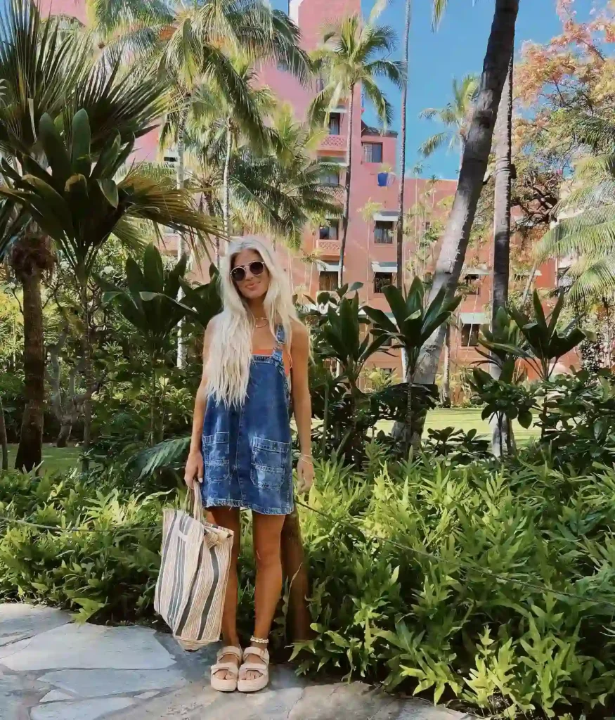 Woman in denim dress with sunglasses standing amidst tropical plants, holding a striped tote bag in a sunny garden.