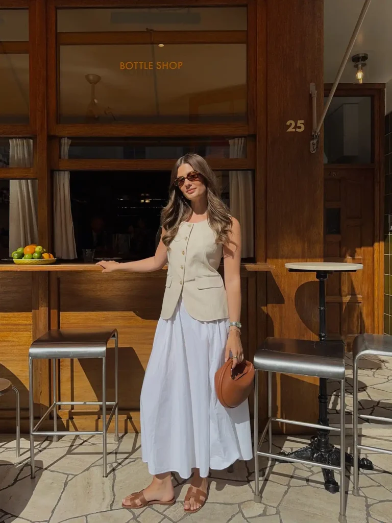 Young woman in a beige vest and white skirt stands outside a bottle shop, enjoying a sunny day with stylish accessories.