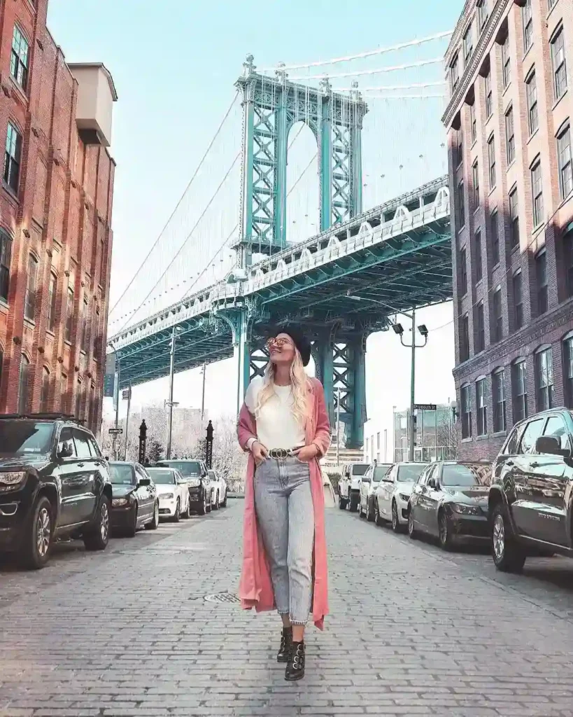 Woman in stylish outfit walking on a cobblestone street with the Manhattan Bridge in the background.