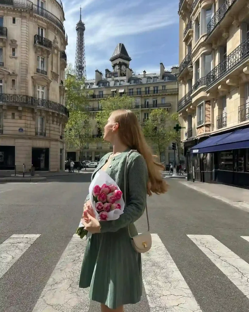 Young woman in a green dress holding pink flowers, standing on a street in Paris with the Eiffel Tower in the background.